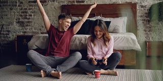 丈夫和妻子坐在家里卧室的地板上拿着操纵杆玩电子游戏，男人赢了，女人输了，两人都在享受游戏。