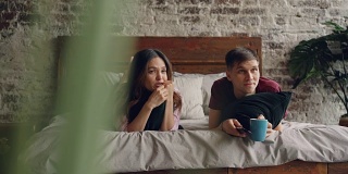 一个年轻人和他的女朋友躺在床上拿着遥控器看电视，他们拿着枕头和杯子。电视、家庭生活和娱乐理念。
