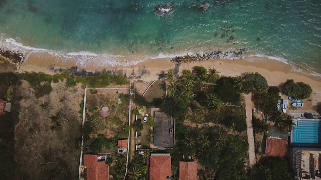 封锁航拍的异国情调的海岸度假屋与棕榈树，海浪冲刷可爱的海滩在一个阳光明媚的日子