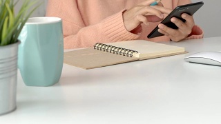 年轻的亚洲女性企业主在办公室使用智能手机应用程序并做笔记。视频素材模板下载