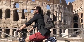 三个年轻的朋友游客在阳光明媚的日子里骑着自行车在罗马市中心的罗马斗牛场周围的道路上慢镜头摄影机汽车稳定摄影机
