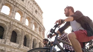三个快乐的年轻朋友，游客骑着自行车背包在罗马斗牛场在阳光明媚的一天慢镜头稳定摄影机地面拍摄视频素材模板下载