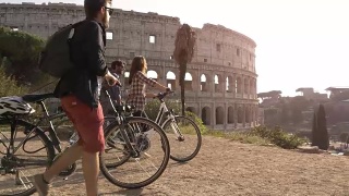 三个快乐的年轻朋友游客与自行车和背包在罗马斗牛场到达山与树木在日落慢镜头摄影机视频素材模板下载