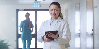 美丽的年轻女医生站在医院走廊，用平板电脑和迷人的微笑。工作中的专业人士。