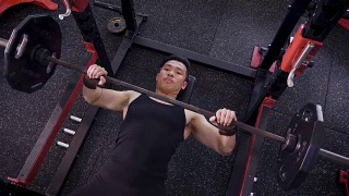 年轻的亚洲肌肉男子躺着和锻炼杠铃卧推在健身房，生活方式运动和健美的概念视频素材模板下载