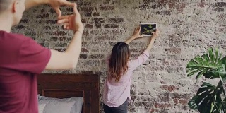 迷人的阿兰女孩正在砖墙上挑选照片的位置，而她的丈夫正在用手指做框架形状，看着她。装修平面的概念。