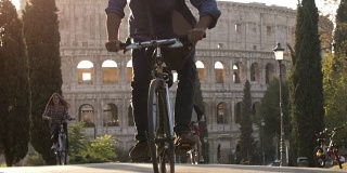 三个年轻的朋友游客骑自行车在colle oppio公园前的罗马斗牛场在路上与树木日落在罗马慢镜头