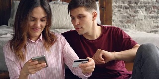 一个漂亮的亚洲女人正在网上购物，用智能手机用银行卡支付，然后拥抱她的丈夫坐在家里的地板上。便捷的网络购物理念。