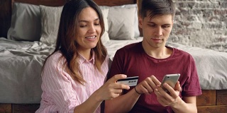 英俊的年轻人正在用智能手机支付网上购物，而他有魅力的妻子正坐在他旁边，拿着银行卡和他聊天。
