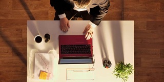 一个戴眼镜穿西装的年轻人坐在桌子后面拿着笔记本电脑和咖啡，准备离开，拍得好