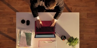 一个戴眼镜穿西装的年轻人走了进来，喝着咖啡，用笔记本电脑工作，坐在桌子后面，上图