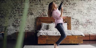 有趣的女孩拿着吹风机在卧室的地毯上跳舞唱歌，靠近双人床。活跃的年轻女子享受自由时间和听音乐。