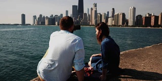 在美国芝加哥的密歇根湖上野餐的年轻夫妇的后视图。男人用智能手机拍照