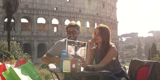 快乐的年轻游客夫妇阅读菜单，选择食物和饮料，坐在罗马斗牛场前的酒吧餐厅在日落准备点菜