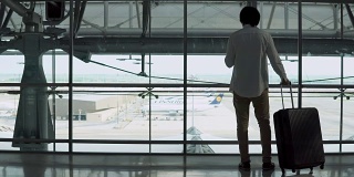 4 k。男性年轻乘客使用智能手机带着行李箱在机场候机楼出发区行走。穿着休闲装出差的亚洲商人。现代旅游生活理念。