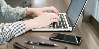 年轻的亚洲商人在工作空间使用笔记本电脑，木桌上放着智能手机和笔记本。男性用手在笔记本电脑键盘上打字。数字时代概念中的自由职业生活方式。