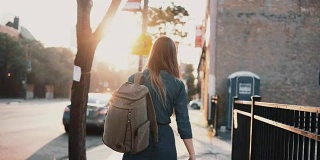 背影年轻时尚的女人与背包独自走在夏天的日落，花时间在市中心