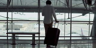 4 k。男性年轻乘客使用智能手机带着行李箱行走，坐在机场候机楼出发区的长凳上。正在出差的亚洲商人。现代旅游生活理念