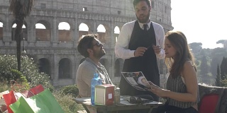 日落时分，在罗马圆形大剧场前的酒吧餐厅里，一对快乐的年轻游客正在点餐和饮料，优雅的服务员正坐在那里