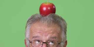 在绿色背景下，一名年长男子头顶苹果的大头照