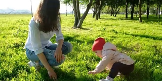 小孩试着站起来，迈出第一步。快乐的年轻母亲和婴儿坐在公园的草地上
