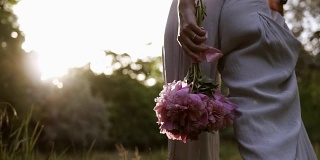 华丽的低角度的观点，一个苗条的女人走在户外与粉红色的牡丹花在手里。阳光照在背景上。腿的目的片段