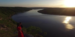 从空中飞离的情侣站在悬崖边靠近水边。穿着红色裙子的女人。人黑。日落