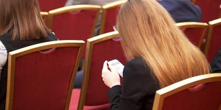在商务会议上，人们坐在大厅里的红椅子上，女性手里拿着智能手机