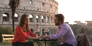 日落时分，浪漫优雅的情侣坐在罗马斗牛场前的餐厅桌前，手里拿着一杯红酒，手拉手交谈