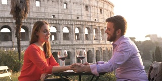 日落时分，浪漫优雅的情侣坐在罗马斗牛场前的餐厅桌前，手里拿着一杯红酒，手拉手交谈