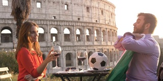日落时分，在罗马罗马圆形大剧场前，一对浪漫的年轻情侣坐在餐厅的餐桌前，一个年轻人和他的女友正在用意大利国旗踢足球