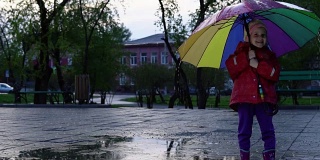 可爱的小女孩拿着五颜六色的雨伞在夕阳下的公园里的水坑里跳跃。