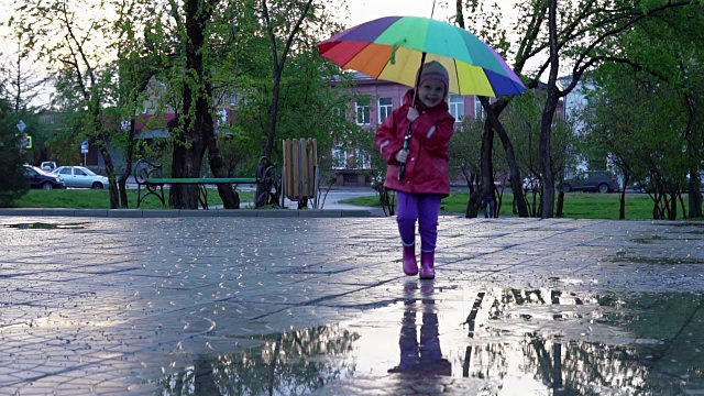 可爱的小女孩拿着五颜六色的雨伞在夕阳下穿过公园的水坑
