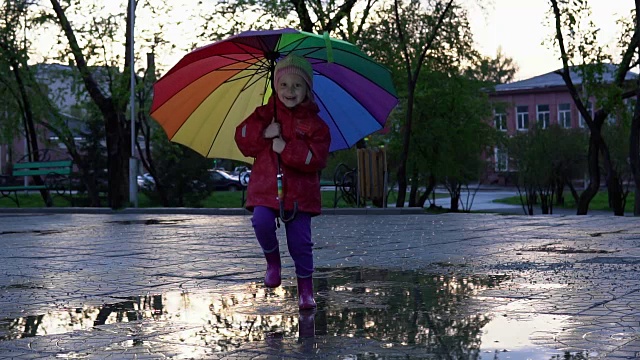 可爱的小女孩拿着五颜六色的雨伞在夕阳下的公园里的水坑里跳跃。