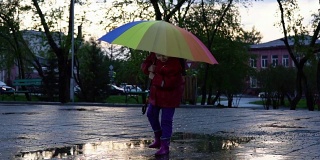 可爱的小女孩拿着五颜六色的雨伞在夕阳下的公园里的水坑里跳跃。
