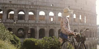 美丽的年轻女子在五颜六色的时装穿着独自走在山上与自行车在罗马斗牛场前在日落与树迷人的女孩草帽