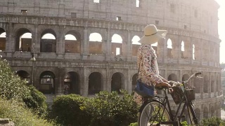 美丽的年轻女子在五颜六色的时装穿着独自走在山上与自行车在罗马斗牛场前在日落与树迷人的女孩草帽视频素材模板下载