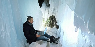 一个人正在冰洞里用笔记本电脑上网。围绕着神秘美丽的冰洞。用户通过社交网络进行交流。他穿着黑色的外套，梳着时髦的发型，是个疯狂的嬉皮士。