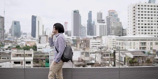 年轻的亚洲商人用智能手机在写字楼屋顶平台上谈生意，背景是城市景色。商务沟通和企业理念。