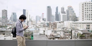 年轻的亚洲商人在办公大楼屋顶露台上使用智能手机，背景是城市景色。在智能设备上使用社交媒体应用程序或玩游戏。