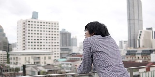 年轻的亚洲商人站在办公楼屋顶的露台上，看着背后的城市景色。城市生活方式的概念。