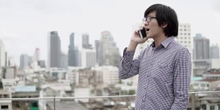 年轻的亚洲商人站在写字楼屋顶露台上用智能手机谈生意，背景是城市景色。商务沟通和企业理念。