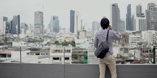 年轻的亚洲商人站在办公楼屋顶露台上，用智能手机谈生意，背景是城市景色。商务沟通和企业理念。