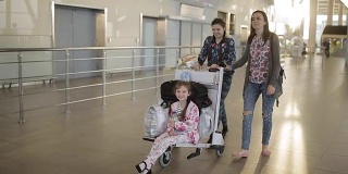 一位女士带着两个女儿，用手推车拖着行李沿着机场大厅走。乘客在等候区。
