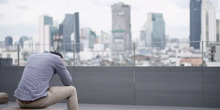 年轻的亚洲商人感到压力和沮丧的工作和生活问题坐在办公大楼屋顶露台的长凳上。背景中的城市视图。重度抑郁症的概念。
