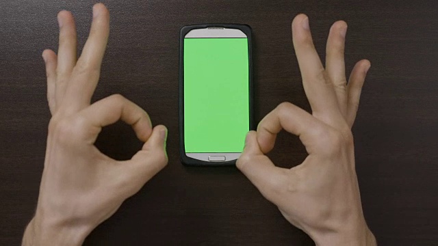 千禧一代的男人在绿色屏幕智能手机前做手势，用大拇指向上OK心的手指手势评估应用程序