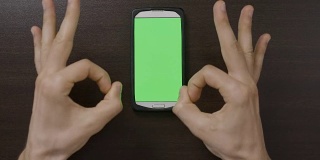 千禧一代的男人在绿色屏幕智能手机前做手势，用大拇指向上OK心的手指手势评估应用程序