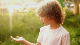一个卷发男孩的闭合肖像。这个男生站在夏雨中，用手擦着雨滴。温暖的雨。儿童一天视频素材模板下载
