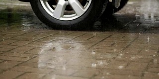 特写一辆车的轮子，车被打湿了在停车场，大滴大滴地溅成水坑，又下起了大雨，倾盆大雨，整个停车场都被水淹没了