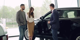 一对有吸引力的夫妇正在与汽车销售经理在豪华汽车经销商和看美丽的黑色汽车。豪华汽车和人的概念。